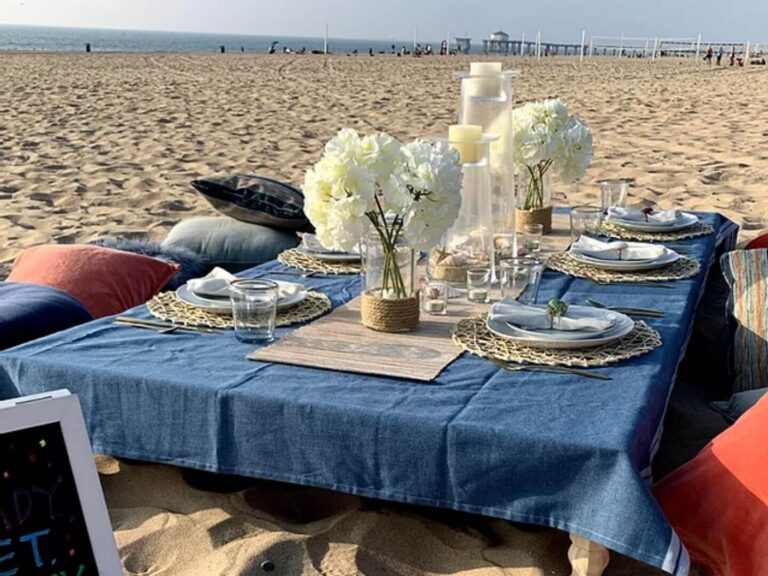 beach picnic set up with a table, table cloth, flowers, plates and Cushions